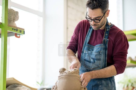 Photo for Portrait of ceramist dressed in an apron working on clay sculpture in bright ceramic workshop. - Royalty Free Image