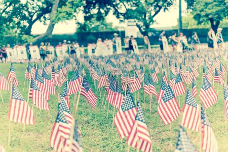 "Filtered image lawn American flags with blurry row of people carry fallen soldiers banners parade"