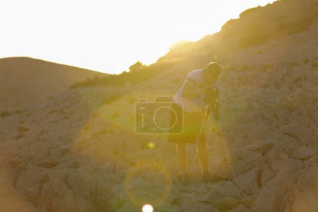 Téléchargez les photos : Photographe installant la caméra pendant le coucher du soleil sur la colline rocheuse - en image libre de droit