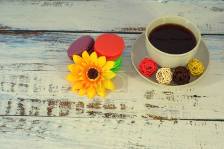Téléchargez les photos : "Quatre macarons de différentes couleurs ornés d'un bouton de fleur sont couchés sur une table à côté d'une tasse de café sur une soucoupe avec une cuillère. Gros plan
." - en image libre de droit