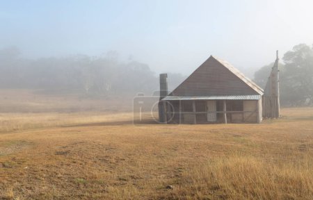 Téléchargez les photos : Cabane en rondins dans les montagnes enneigées avec brouillard tôt le matin - en image libre de droit