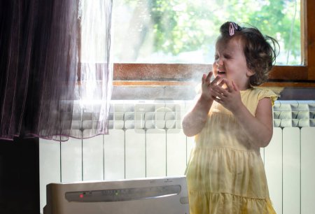 Téléchargez les photos : Petite fille dans une pièce poussiéreuse. Purificateur d'air et toux enfant - en image libre de droit