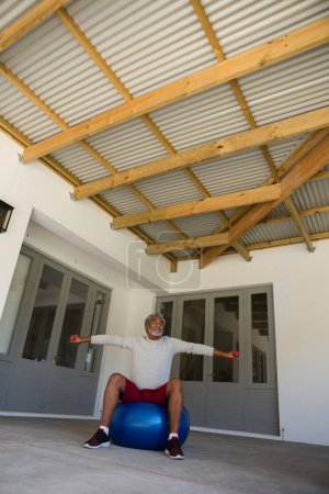 Foto de Hombre mayor haciendo ejercicio con pesas en la pelota de ejercicio en el porche - Imagen libre de derechos