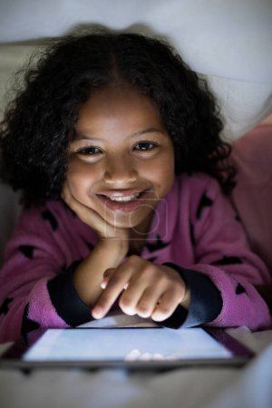 Téléchargez les photos : Souriant fille en utilisant tablette numérique sous couverture dans la chambre - en image libre de droit