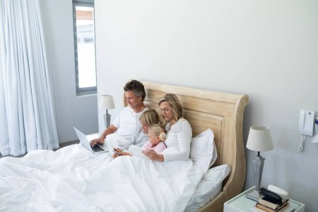 Téléchargez les photos : Famille utilisant tablette numérique, téléphone portable et ordinateur portable sur le lit dans la chambre - en image libre de droit