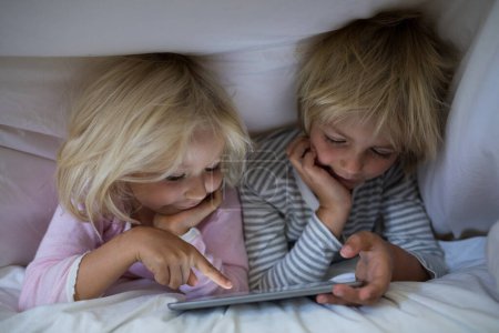 Téléchargez les photos : Frères et sœurs utilisant une tablette numérique sous la couverture sur le lit dans la chambre - en image libre de droit
