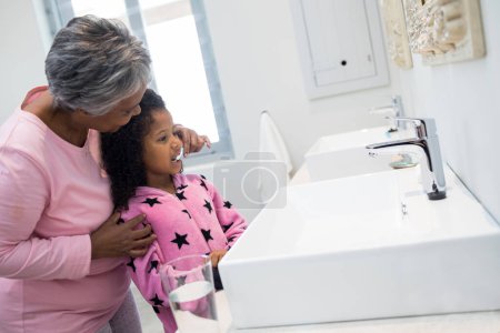 Téléchargez les photos : Grand-mère aidant sa petite-fille à se brosser les dents dans la salle de bain - en image libre de droit
