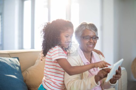 Téléchargez les photos : Grand-mère et petite-fille souriantes utilisant une tablette numérique sur le canapé dans le salon - en image libre de droit