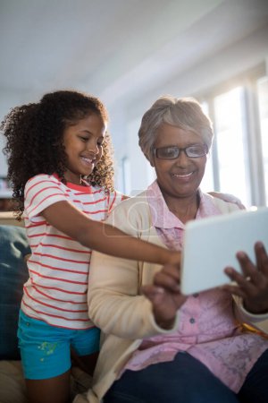 Téléchargez les photos : Grand-mère et petite-fille souriantes utilisant une tablette numérique sur le canapé dans le salon - en image libre de droit