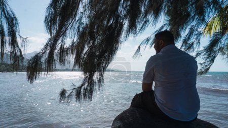 Téléchargez les photos : Avec l'homme assis sur un rocher relaxant et réfléchissant avec un beau ciel bleu et la mer - en image libre de droit