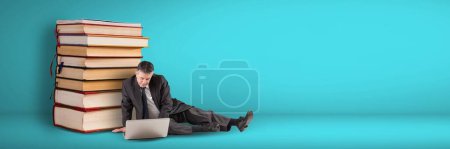 Téléchargez les photos : Homme d'affaires utilisant un ordinateur sur le sol à côté d'une pile de livres sur fond bleu - en image libre de droit