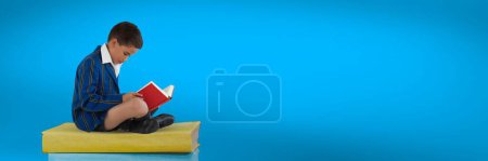 Photo for Boy reading and sitting on a pile of books with blue background - Royalty Free Image