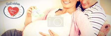 Foto de Imagen compuesta del retrato de una mujer embarazada feliz sosteniendo zapatos de bebé y de su marido en un sofá - Imagen libre de derechos