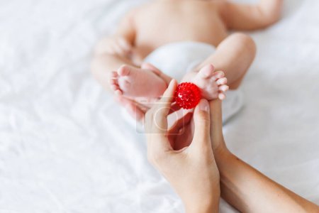 Téléchargez les photos : Mère tient les pieds du nouveau-né. Petits doigts et boule de massage rouge dans la main de la femme. Matin confortable à la maison - en image libre de droit