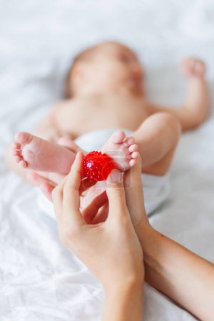 Téléchargez les photos : Mère tient les pieds du nouveau-né. Petits doigts et boule de massage rouge - en image libre de droit