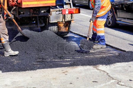 The working crew evenly distributes hot asphalt with shovels manually on the repaired site of the road.