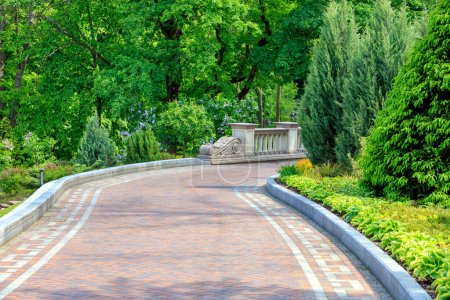 Photo for "Walkway paved with tiles in a beautiful park, framed by different bushes and flowers, in soft rays of morning light." - Royalty Free Image
