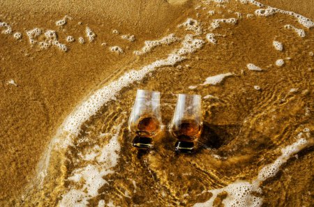 Foto de Dos vasos de whisky de malta en la arena lavada por la ola - Imagen libre de derechos