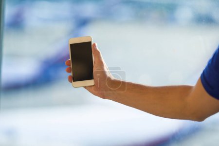 Téléchargez les photos : "Gros plan de jeune homme utiliser le téléphone intelligent dans la fenêtre arrière-plan de l'aéroport
" - en image libre de droit