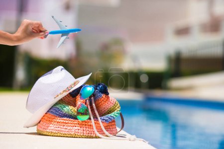 Foto de Colorido bolso de playa, sombrero de paja, gafas de sol y modelo de avión en la mano cerca de la piscina - Imagen libre de derechos