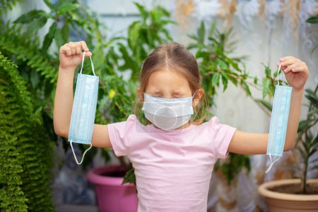Photo for A little girl in a medical mask holds another mask and cries. Tired of isolation. - Royalty Free Image