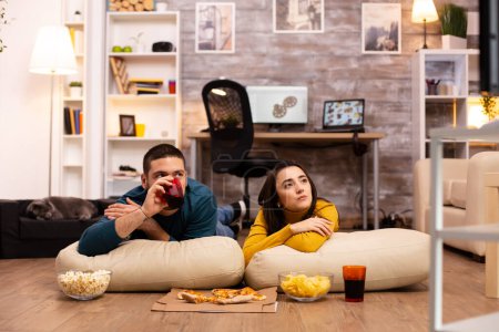 Téléchargez les photos : Couple assis sur le sol et regarder la télévision dans leur salon - en image libre de droit