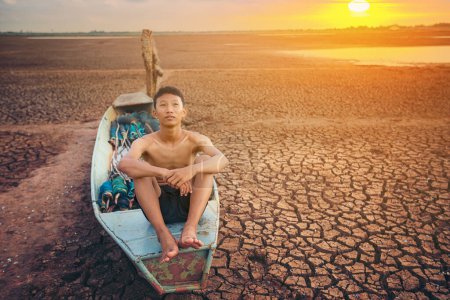 Téléchargez les photos : Triste garçon assis sur un bateau qui était garé dans un sol aride - en image libre de droit
