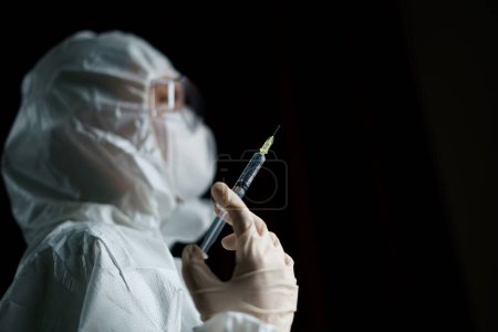 Photo for Woman wearing gloves with biohazard protective suit and mask holding syringe - Royalty Free Image