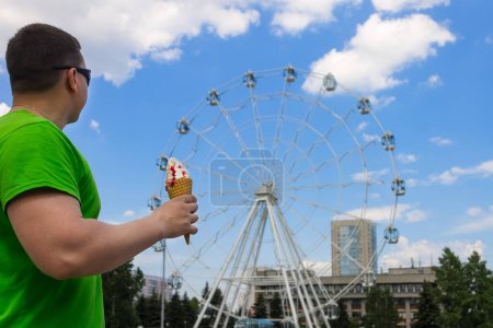 Foto de Un hombre con un helado en la mano está de pie en un parque de diversiones mirando una noria - Imagen libre de derechos
