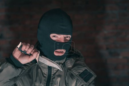 Photo for A thug in a balaclava with a knife against a brick wall in the dark shows threatening gestures - Royalty Free Image