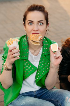 Foto de Chicas divertidas comiendo hamburguesa y patatas fritas en la calle - Imagen libre de derechos
