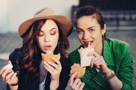 Foto de Chicas divertidas comiendo hamburguesa y patatas fritas en la calle - Imagen libre de derechos