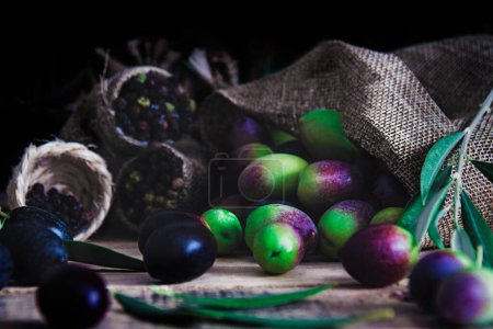 Téléchargez les photos : Nature morte d'olives noires, vertes et violettes en sac et avec des feuilles d'olivier. Technique alimentaire sombre et concept alimentaire - en image libre de droit