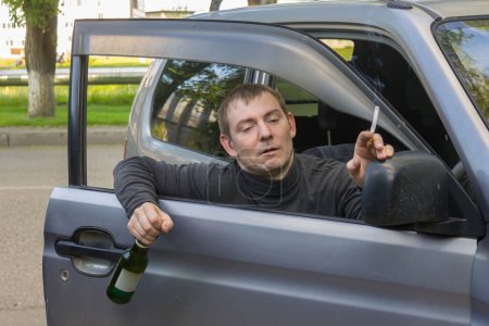 Téléchargez les photos : La conduite en état d'ivresse. Pilote ivre accroché à la porte de sa voiture avec une bouteille de bière et une cigarette - en image libre de droit