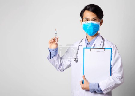 Photo for "asia man doctor wearing surgical mask and hand holding vaccine syringe and paper form standing in gray wall background. Concept Preventive measures against corona virus." - Royalty Free Image