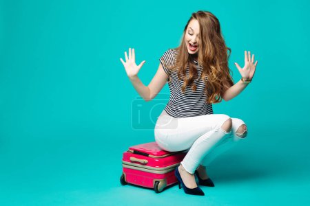 Téléchargez les photos : Émotionnellement fille assise sur un sac de voyage et gesticulant émotionnellement - en image libre de droit