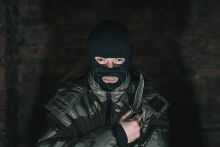 Foto de Peligroso criminal en una máscara con un cuchillo en la oscuridad contra una pared de ladrillo - Imagen libre de derechos