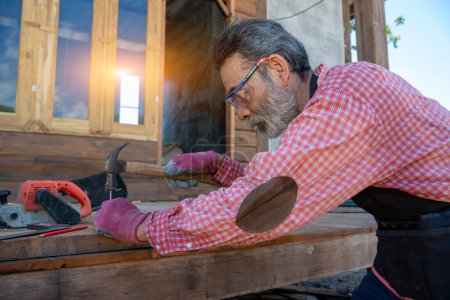 Téléchargez les photos : Menuisier sénior à l'aide du marteau frappé un clou pour l'assemblage du bois - en image libre de droit