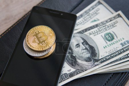 Photo for "Golden bitcoins on the smart phone and us dollars with notebook on background. Bitcoin crypto currebcy. Digital currency. Profit from mining crypto currencies. Miner with dollars from trading." - Royalty Free Image