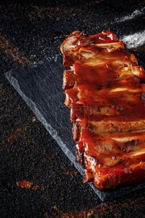 Foto de Primer plano de costillas de cerdo al horno con salsa barbacoa en el fondo de pizarra, con patatas y especias al horno, pan y cubiertos - Imagen libre de derechos