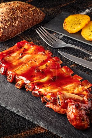 Foto de Primer plano de costillas de cerdo al horno con salsa barbacoa en el fondo de pizarra, con patatas y especias al horno, pan y cubiertos - Imagen libre de derechos