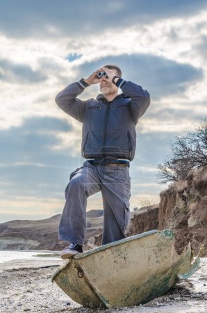 Photo for Man stands in a broken boat on the beach and carefully looks through binoculars - Royalty Free Image