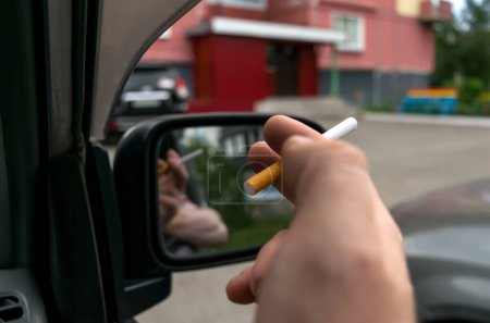 Téléchargez les photos : Gros plan d'une cigarette dans la main d'un homme dans la voiture, qui regarde la porte d'entrée de la maison - en image libre de droit