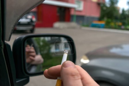 Téléchargez les photos : Gros plan d'une cigarette dans la main d'un homme dans la voiture, qui regarde la porte d'entrée de la maison - en image libre de droit