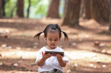 Photo for Asian little child girl sitting in the garden and using smartphone to look cartoon with earphone. - Royalty Free Image