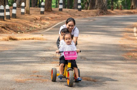 Foto de Asiática joven madre sosteniendo bicicleta a sus hijas, que montar en bicicleta con su hermana en el camino en el jardín. - Imagen libre de derechos
