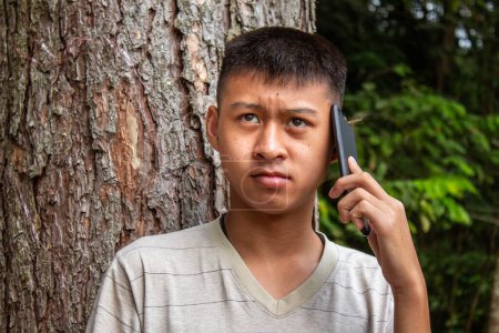 Photo for Asian teen boy standing in the garden and talking on phone seriously. - Royalty Free Image
