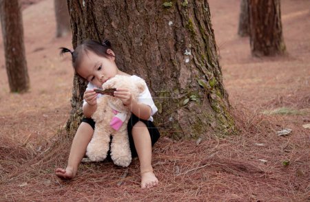 Téléchargez les photos : Asiatique petite fille enfant est étreignant ours et assis sur le sol sous le pin seul - en image libre de droit