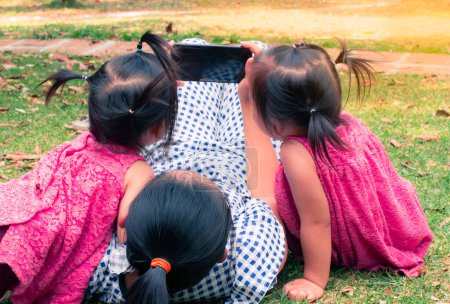 Photo for Asian mother and her daughters sitting on the grasses ground in the garden and looking at smartphone happily - Royalty Free Image