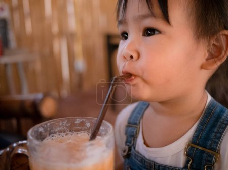 Téléchargez les photos : Joyeux petit asiatique fille manger thai thé glacé avec du lait dans le verre à l'extérieur du café - en image libre de droit